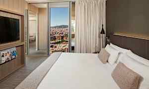 Ett hotellrum med en stor säng med vita sängkläder och beiga kuddar, en modern design med ett TV-skåp, tidskrifter och ett fönster som visar en stadsvy. (AI-genererad text)