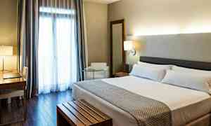 Ett hotellrum med en dubbelsäng, ett skrivbord och stol, en fåtölj, och ett stort fönster med gardiner. (AI-genererad text)