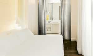 Ett hotellrum med en vit säng och ett modernt badrum synligt genom vertikala svarta paneler. (AI-genererad text)