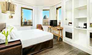 Ett hotellrum med en dubbelsäng, mörka trägolv och en skrivbord med TV. Rummet har stora fönster som visar en fin utsikt över staden. (AI-genererad text)