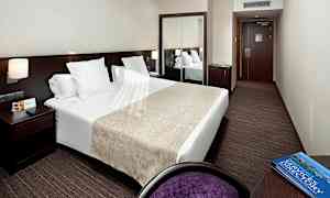 Ett hotellrum med en dubbelsäng, sängbord med lampa, spegel och en skrivbordsstol. (AI-genererad text)