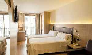 Ett hotellrum med två enkelsängar, ett skrivbord och en stol framför en stor fönster med balkongräcke. Ett TV är monterad på väggen. (AI-genererad text)