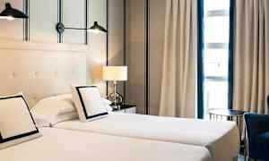 Två enkelsängar står bredvid varandra med kuddar och vit sängklädsel. Bakom sängarna finns en ljus vägg med randmönster. Intill sängen står ett bord med en bordslampa och ett fönster med gardiner syns i bakgrunden. (AI-genererad text)