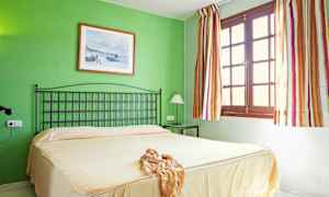 Ett sovrum med en grön vägg, en säng med beige överkast, en svart sänggavel, och ett fönster med randiga gardiner. (AI-genererad text)