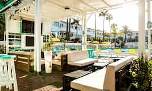 En utomhuslounge under en vit pergola med möbler och växter, med utsikt över en pool och omgivande byggnader. (AI-genererad text)