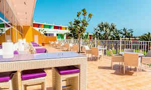 En utomhusterrass med bord och stolar, omgiven av palmer och färgglada byggnader. (AI-genererad text)
