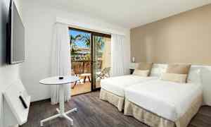 Ett modernt hotellrum med två enkelsängar, ett mindre bord och en väggmonterad TV. Rummet har ljusa färger och ett fönster som leder ut till en balkong med utsikt mot palmer. (AI-genererad text)