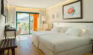 Ett ljust och modernt hotellrum med två sängar, en sittgrupp och ett skrivbord. En balkong med utsikt över landskapet syns i bakgrunden. (AI-genererad text)