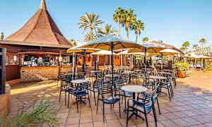 En utomhusrestaurang med bord, stolar och parasoller. Det finns en bar med ett tak i bakgrunden samt omgivande palmer. (AI-genererad text)