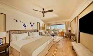 Ett hotellrum med en dubbelsäng, sittmöbler, ett skrivbord och en TV. Rummet har en modern inredning med blå dekorationer på väggen och ett fönster som leder till en balkong med utsikt. (AI-genererad text)