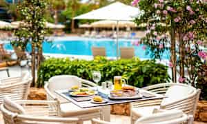 Ett bord med mat och dryck står på en uteservering nära en pool omgiven av blommande växter och parasoller. (AI-genererad text)