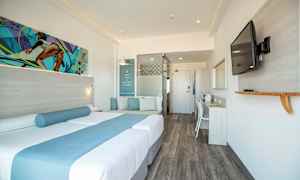 Ett modernt hotellrum med en dubbelsäng dekorerad med blå textilier, en sittplats med kuddar, ett skrivbord, och en väggmonterad TV. (AI-genererad text)