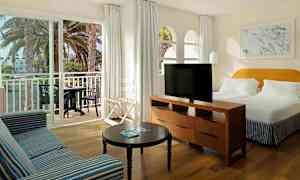 Bild av ett hotellrum med en stor säng, en soffa, ett bord och en TV. En balkong med bord och stolar syns genom skjutdörrar. (AI-genererad text)