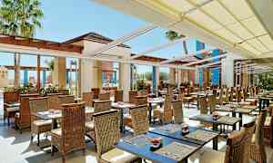 En utomhusrestaurang med många bord och stolar under en pergola med tak. Palmtäd syns i bakgrunden. (AI-genererad text)