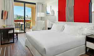 Ett hotellrum med en stor säng, en fåtölj, ett bord med stolar, och en balkong med utsikt mot en trädgård. (AI-genererad text)