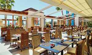 En utomhusrestaurang med sittplatser bestående av bord med stolar i rotting. Solskydd täcker taket och palmer kan ses i bakgrunden. (AI-genererad text)
