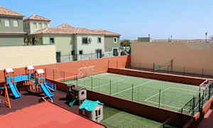Bilden visar en utomhuslekplats med rutschkana och lekhus samt en grön inhägnad sportplan för fotboll och volleyboll. I bakgrunden syns bostadshus. (AI-genererad text)
