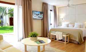 Ett ljust hotellrum med en stor säng, väggmonterad TV, och en sittgrupp framför en rund bord. Stora gardiner med utsikt mot en trädgård. (AI-genererad text)