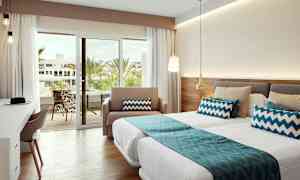 Ett välbelyst hotellrum med sängar, blå dekorativa kuddar och en glasdörr som leder till en balkong med utsikt över palmer. (AI-genererad text)