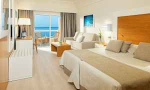 Ett hotellrum med dubbelsäng, sittgrupp och balkong med havsutsikt. (AI-genererad text)