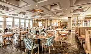 Ett elegant restaurangområde med runda bord och mönstrade stolar, upplyst av taklampor och naturbelyst genom stora fönster. (AI-genererad text)