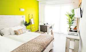 Ett ljust och modernt sovrum med grön accentvägg, säng med vit och beige bäddning, samt bord och stolar nära ett stort fönster. (AI-genererad text)