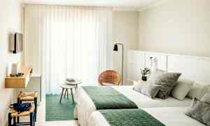 Ett hotellrum med ljusa väggar, två sängar med gröna täcken, en liten sittgrupp med stolar och bord samt en väggmonterad TV. (AI-genererad text)