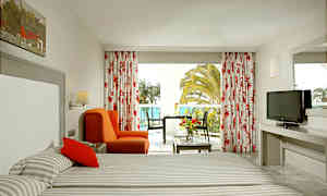 Ett hotellrum med två sängar, en orange fåtölj, ett bord med blommor samt en TV. Fönstret visar utsikt över palmer och havet. (AI-genererad text)