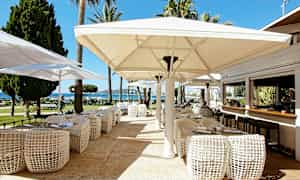 Uteservering med bord, stolar och stora parasoller, med utsikt mot trädgård och hav. (AI-genererad text)