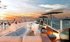 Takbar med utsikt över havet vid solnedgången, med modern möblering inklusive soffor, parasoller och bord. (AI-genererad text)