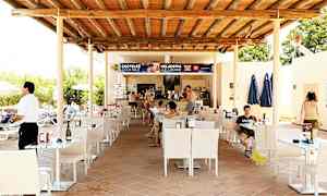 En utomhusservering vid en bar med tak av trä och bord samt stolar. Människor sitter vid borden och beställer från baren. (AI-genererad text)