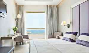 Ett hotellrum med modern inredning. Sängen är bäddad med vita och lila kuddar, och ett stort fönster med utsikt över havet visas. (AI-genererad text)