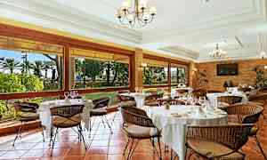 En elegant restaurang med dukade bord och flätade stolar. Stora fönster visar en utsikt över en trädgård. (AI-genererad text)