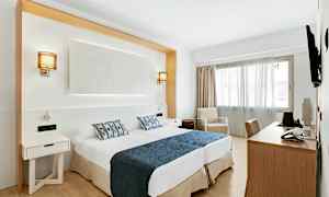 Ett hotellrum med två sängar försedda med blå överkast och mönstrade kuddar, skrivbord, stol och en fåtölj bredvid ett fönster. (AI-genererad text)