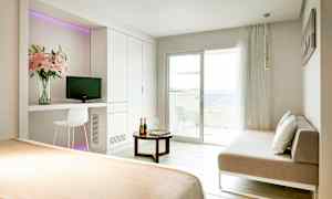 Ett ljust hotellrum med en säng, en soffa, ett bord och en balkong som vetter mot havet. (AI-genererad text)