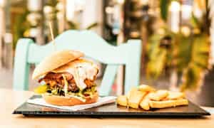 En hamburgare med tillbehör av pommes frites på en tallrik, placerad på ett bord med en stol i bakgrunden. (AI-genererad text)