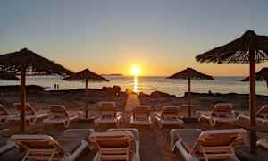 Solsängar på en strand med halmtakparaplyer i förgrunden och en solnedgång vid havet i bakgrunden. (AI-genererad text)