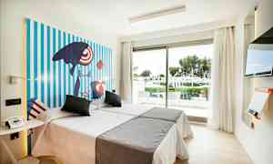 Ett ljust hotellrum med två bäddar, färgglad konst på väggen, en glasdörr med utsikt över en grönskande trädgård och en modern TV. (AI-genererad text)