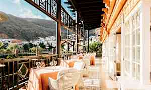 Restaurang med bord och stolar på en terrass med utsikt över byggnader och berg. (AI-genererad text)