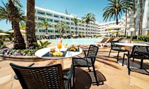 En utomhusmiljö vid en pool med solstolar och parasoller. Ett bord med mat och dryck på sin yta står i förgrunden. (AI-genererad text)