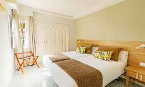 Ett hotellrum med två enkelsängar, dekorativa kuddar och ett brunt överkast, samt en garderob och en stol vid fönstret. (AI-genererad text)