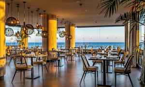 Bild av en restaurang med rottingstolar, borden är dukade med utsikt över havet genom stora glasfönster. (AI-genererad text)