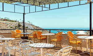 Uteservering med bord och stolar på en terrasse med utsikt över havet och en sluttning med byggnader. (AI-genererad text)