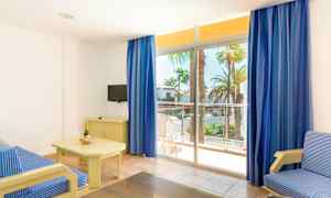 Ett ljust hotellrum med en soffa i blå randigt tyg, ett bord med en champagneflaska och glas, en liten TV på en möbel samt stora fönster med blå gardiner som leder ut mot en balkong med utsikt över palmer. (AI-genererad text)