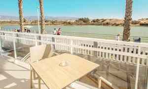 Utsikt från en terrass med bord och stol, med vatten och gångväg i bakgrunden. (AI-genererad text)