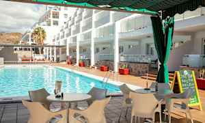 Vy över en swimmingpool med solstolar och bord på terrassen, omgiven av byggnader. (AI-genererad text)