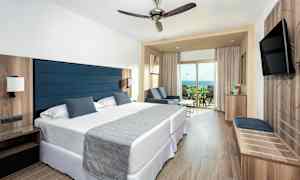 Ett hotellrum med två sängar, ett sittområde och en balkong som har utsikt över havet. (AI-genererad text)