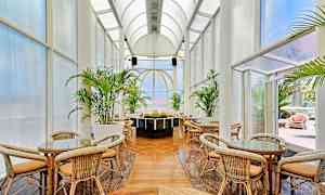 Rostmöbler och bord under högt valvat tak med stora glaspartier omgiven av gröna växter. (AI-genererad text)