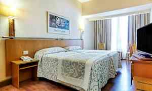 Ett hotellrum med en dubbelsäng med mönstrade överkast, lampor på väggen och en TV på ett skrivbord. Sittmöbler och ett fönster med gardiner finns också. (AI-genererad text)