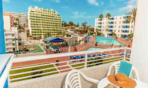 Utsikt från en balkong som visar ett hotellområde med pool och solstolar samt byggnader i bakgrunden. (AI-genererad text)